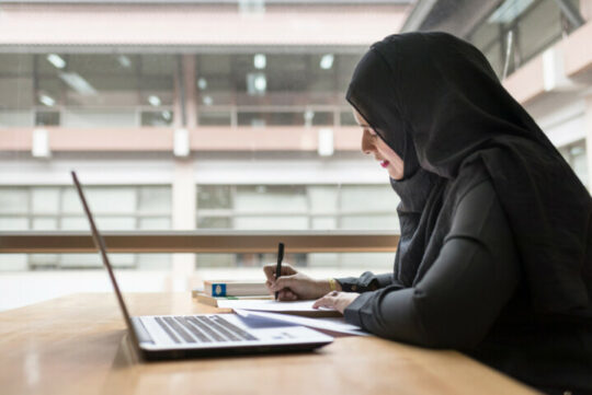 Lady wearing Hijab whilst working on laptop and writing notes on paper