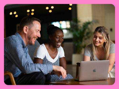 Image shows three people sitting at a table looking at something on an open laptop computer