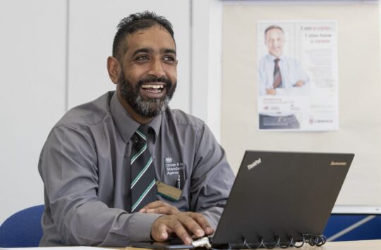 Male DVSA staff member sitting at desk with laptop. He is mid-laugh.