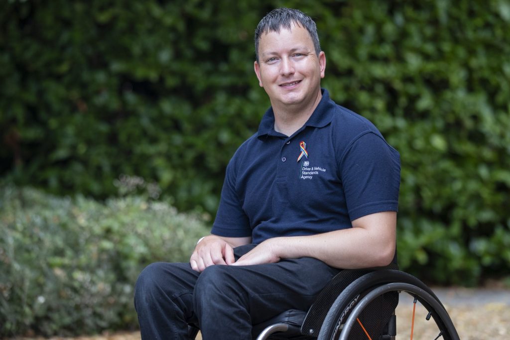 Driving examiner, Nick Kimber. Nick is a wheelchair user and is wearing a rainbow coloured ribbon pinned to his polo shirt. His polo shirt has the Driver & Vehicle Standards Agency logo on it.