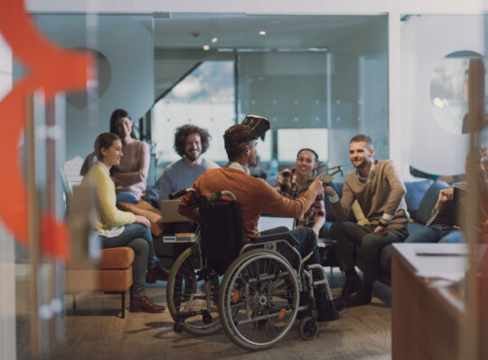 Disabled businessman in a wheelchair at work in modern open space coworking office on team meeting using virtual reality goggles and drone assistance simulation or brainstorming. Effective teamwork of volunteers concept in a startup business.