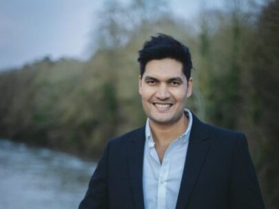 HMRC employee, Akhil, facing the camera and smiling. There is a blurred background of trees and a river