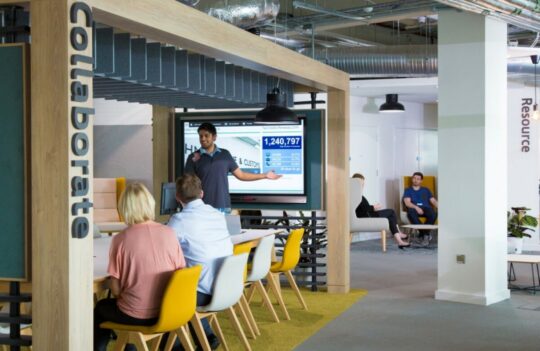 Group of people sitting at a large table in an office, looking at a white board. There is a man standing beside the whiteboard giving a presentation