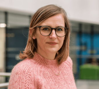 Decorative image: a photo of Anna Tyler. She is wearing a light pink knitted sweater and dark-framed glasses.