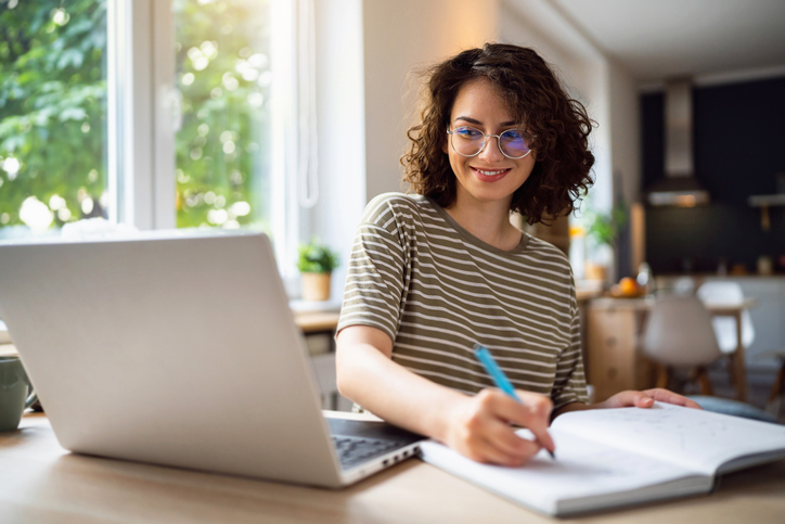 A woman sitting at a laptop is writing in a notepad