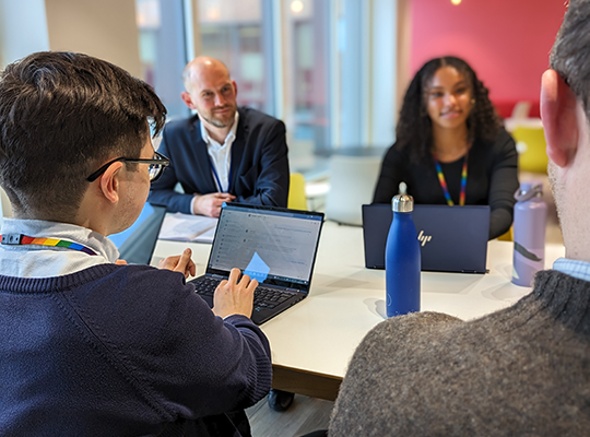 A photo of apprentices around a table, chatting to other team members