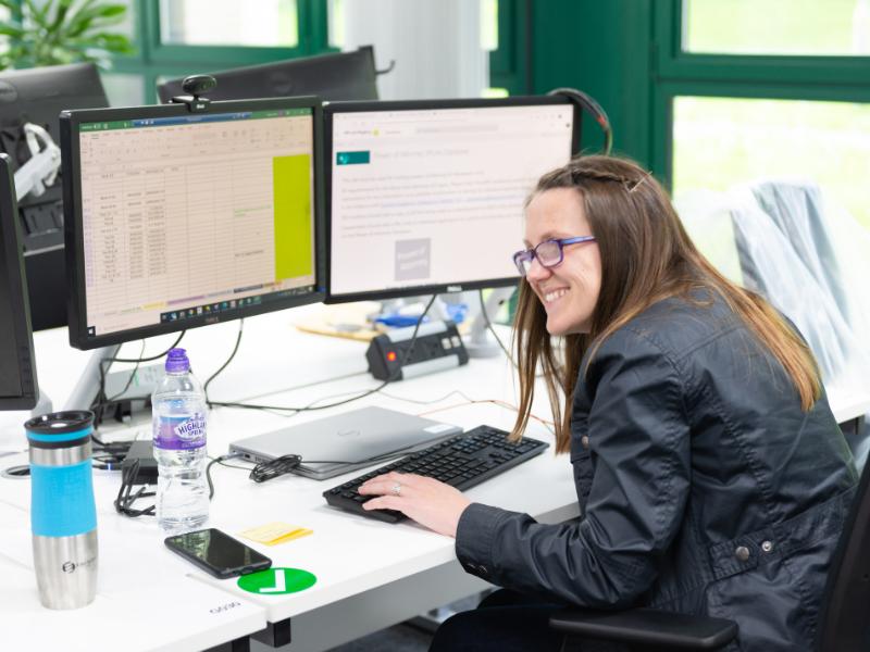 Image of an employee smiling while sitting a desk with 2 screens.