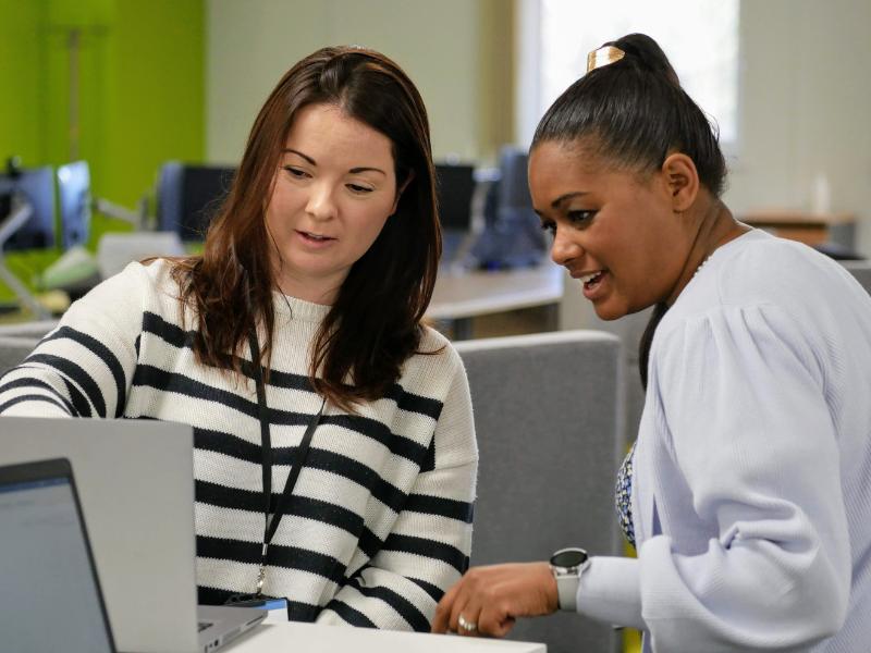 Photo of 2 colleagues in an open plan office discussing something that they are both looking at on a laptop screen.