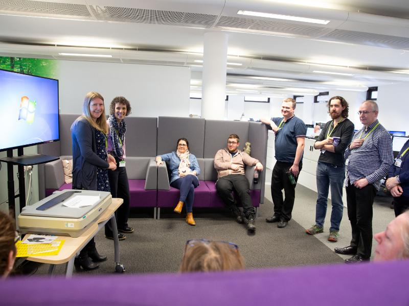 Photo of a group of HM Land Registry Colleagues - in an informal office setting. Some are standing and others are sitting on a grey sofa. They all facing each other and appear to be having a group discussion