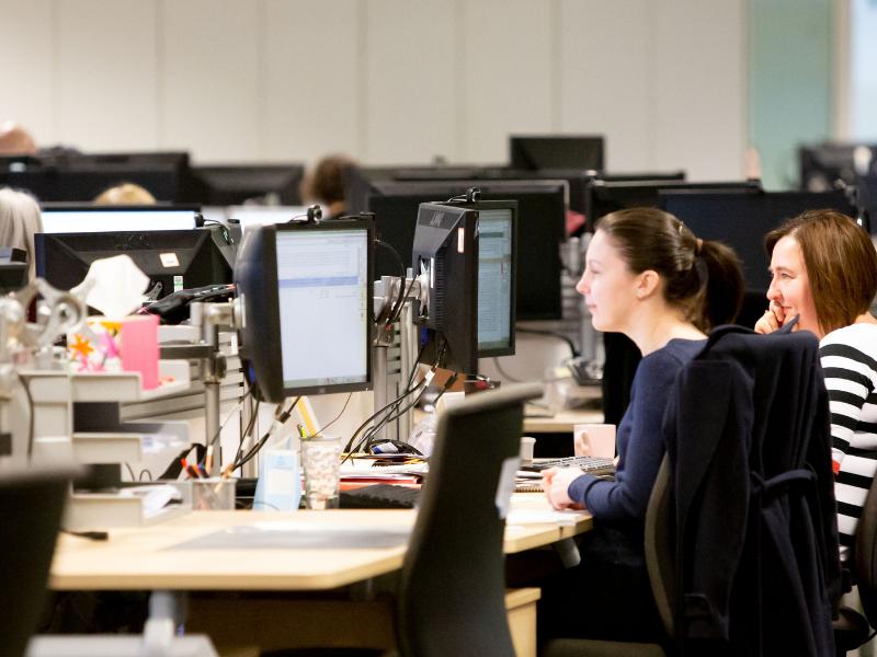 Photo of an open plan office, with 2 employees in the foreground looking at a screen