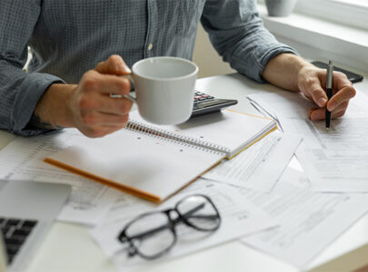 Decorative image: A man from the chest down, sitting at a desk covered with stationery and paperwork, with a coffee cup in his hand.