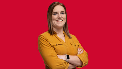 DWP colleague smiling at the camera. Her arms are folded. She is stood against a red background.