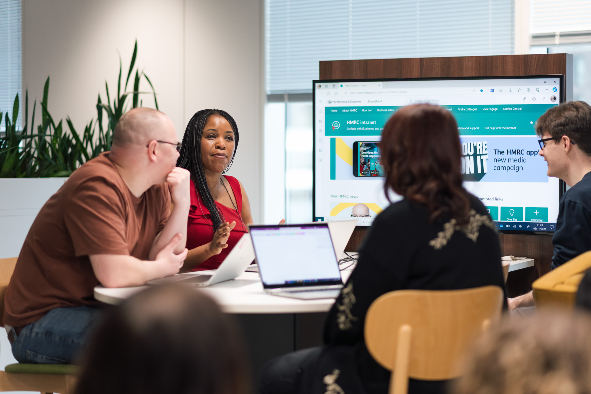 Image of a group of HMRC colleagues sat around a desk in front of a large screen and a laptop.