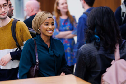 Decorative image: a woman wearing a hijab and teal blouse, speaking to another person at a networking event. The other person is facing away from the camera,