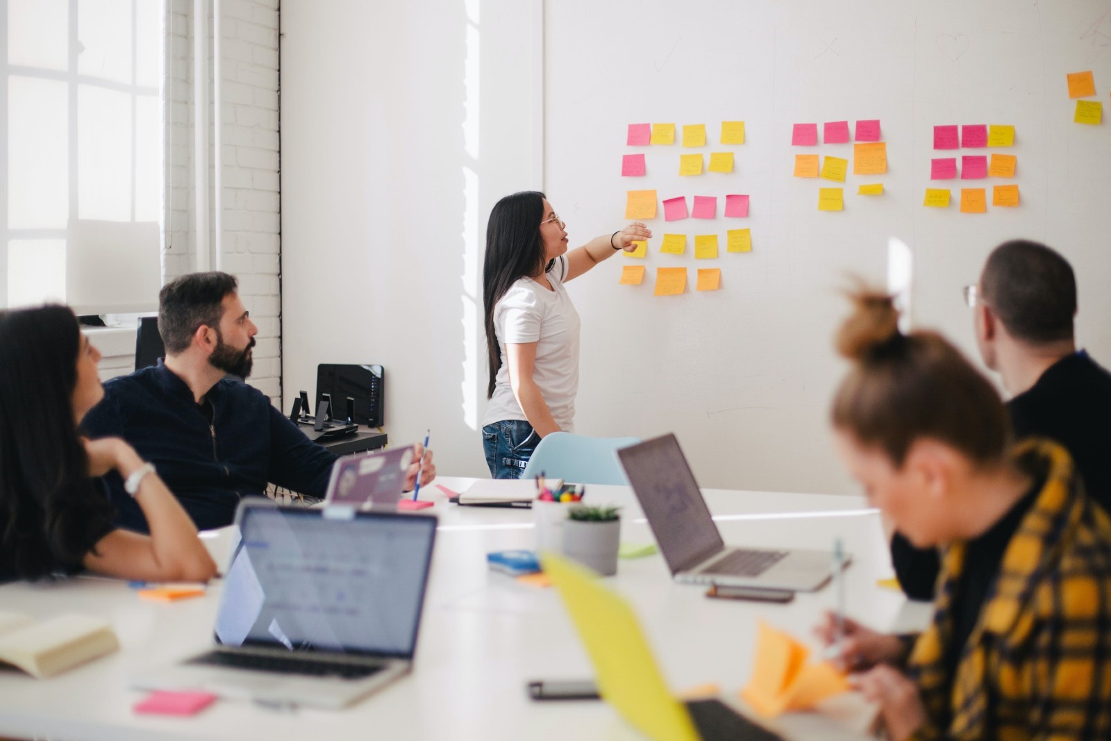 A group of people in an office meeting room with laptops. They are looking at a whiteboard with post it notes and a woman is presenting