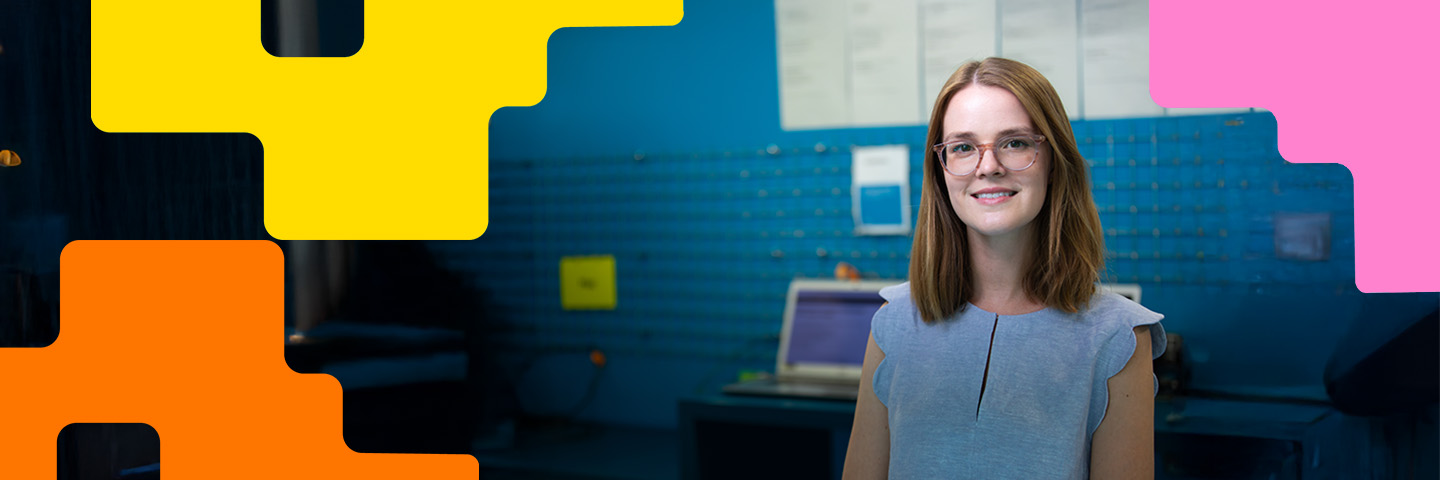 A civil servant smiling with glasses and shoulder-length brown hair looks directly at the camera, wearing a light blue top. They are standing in a digital lab with a blue wall, a laptop, and pinned documents in the blurred background. The panoramic banner is framed by bright yellow, orange, and pink abstract geometric overlays.