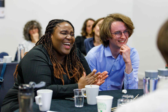 Decorative image: Two colleagues sitting at a table at an event, they are smiling and clapping