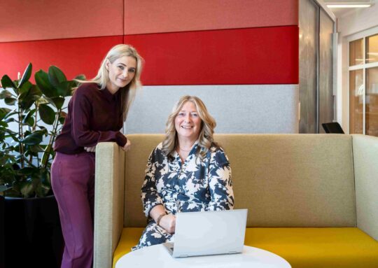 woman sat at table with laptop, female colleague stood beside her, both looking at camera smiling