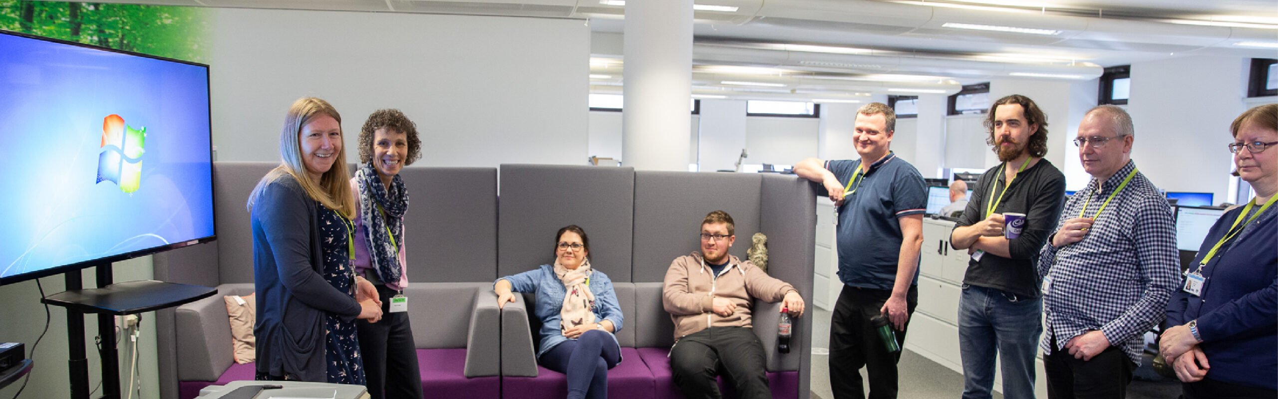 Photo of a number of HM Land Registry colleagues, in an informal setting. Some are standing and others are sitting on a grey sofa. They appear to be discussing something in a group.