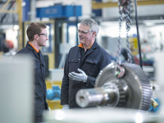 An MCA engineer and apprentice in discussion next to gear wheel.