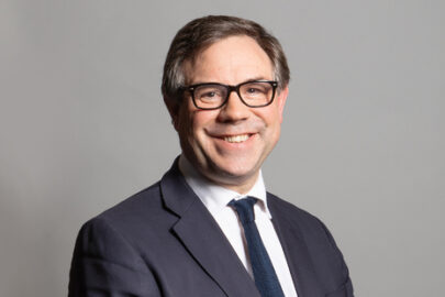 Jeremy Quinn MP wears a suit and smiles at the camera with a grey background