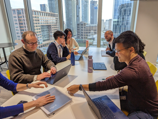 A group of civil servants sitting at a table, working on their laptops, with a city skyline visible through the large windows behind them.