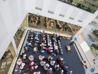 An overhead, top-down view of a large room filled with rows of people sitting down looking towards a person presenting to them. The seated audience are holding information booklets. 