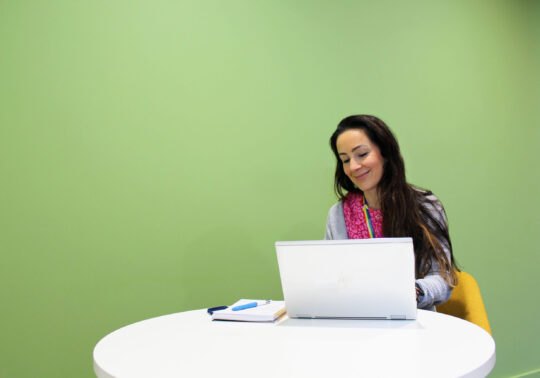 A woman with brown hair smiles whilst working on a laptop against a green wall