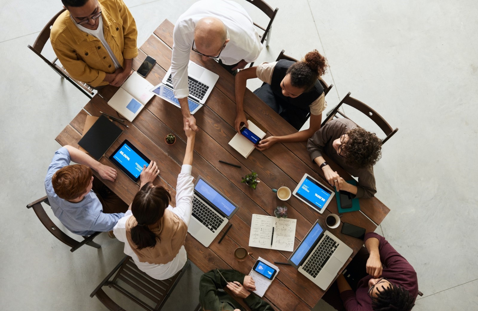 Birds eye view of a group of colleagues sitting at a table with laptops and digital devices. Two people are shaking hands