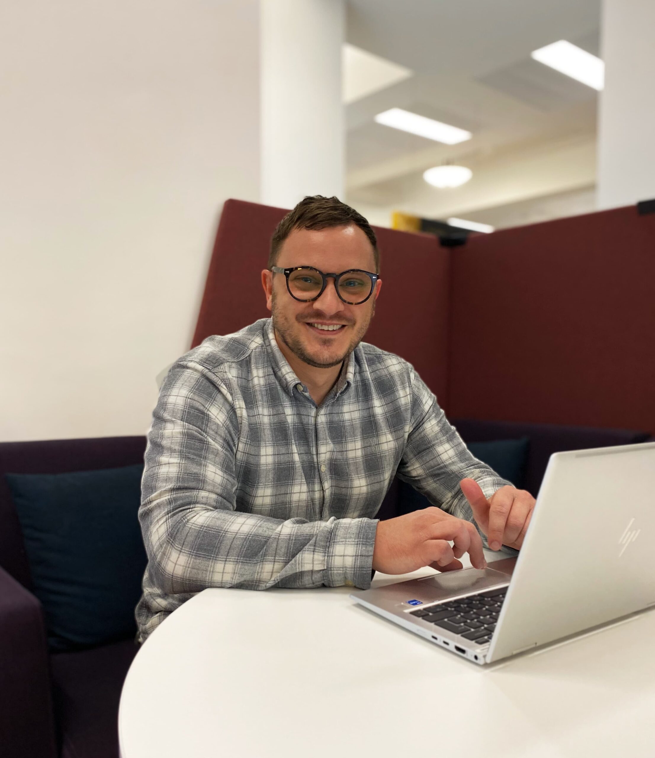 man sat at desk working on laptop, smiling at camera