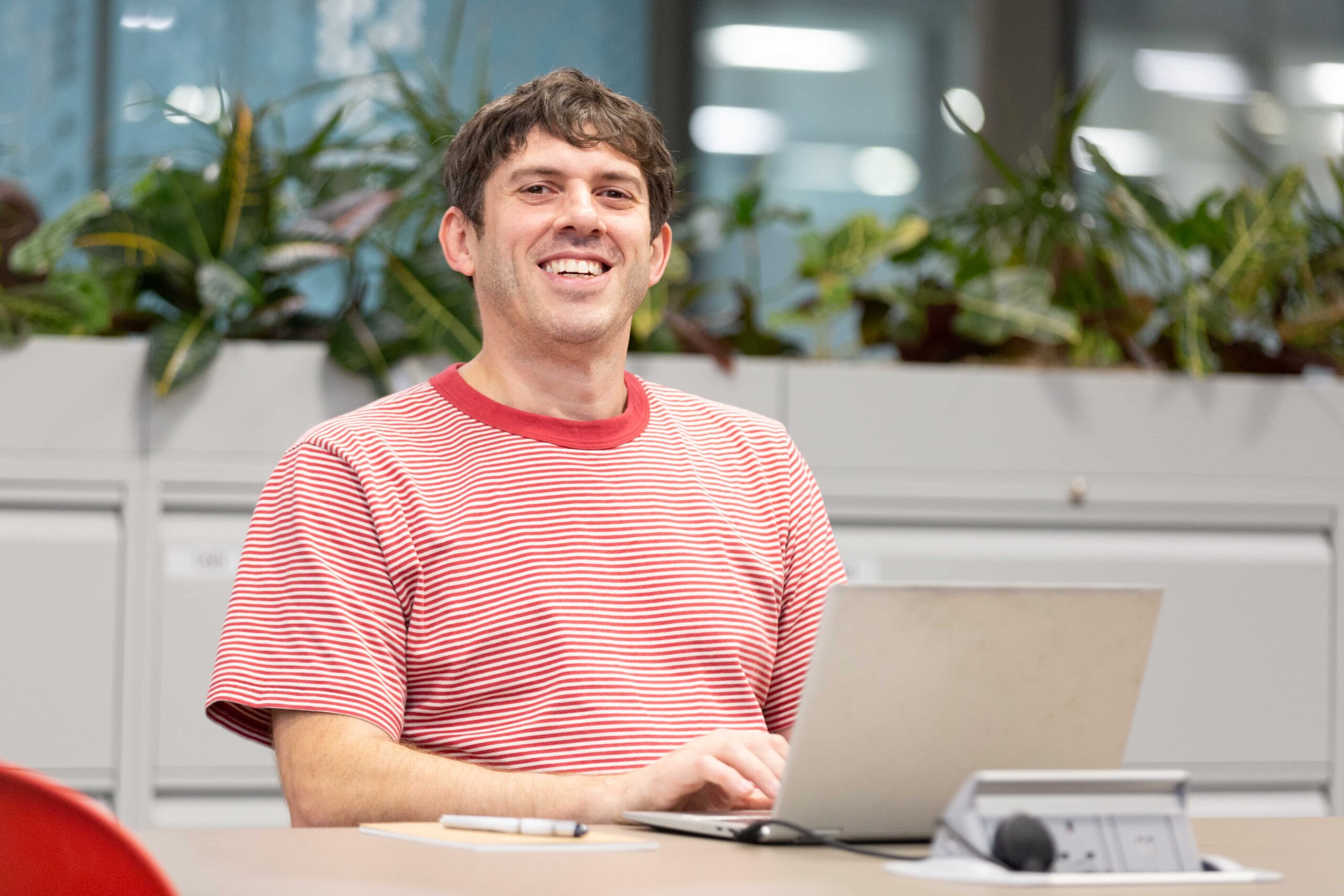 colleague wearing red striped t-shirt sat smiling while working on laptop