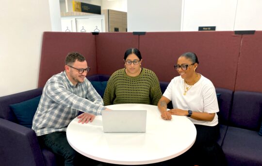 Three colleagues sat round a table, one is pointing at something on a laptop