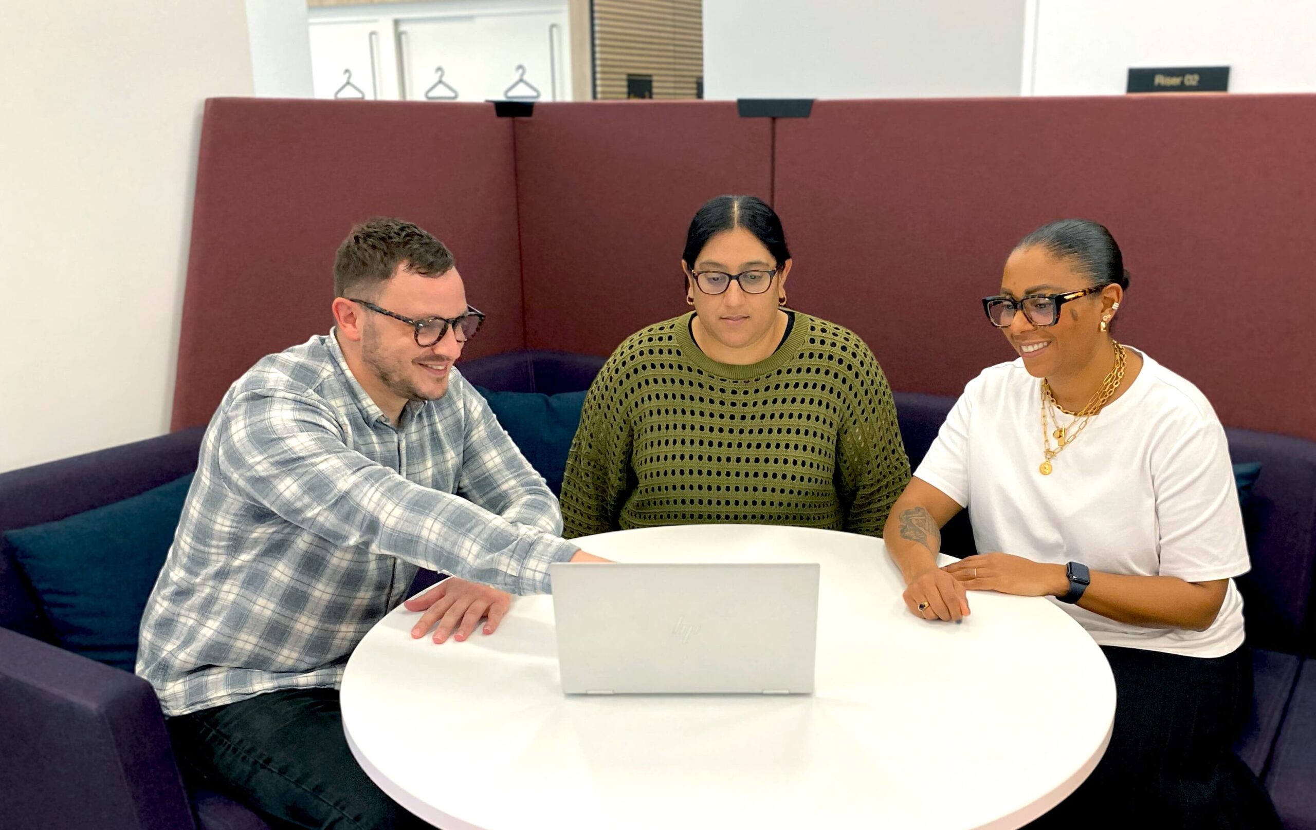 Three colleagues sat round a table, one is pointing at something on a laptop