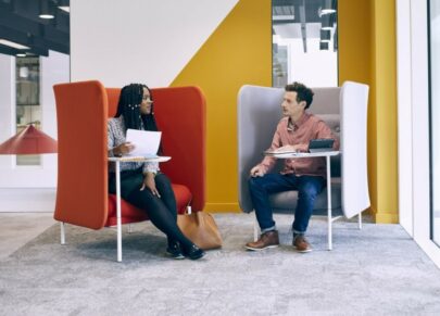 Two people sitting in an office space with laptops, chatting