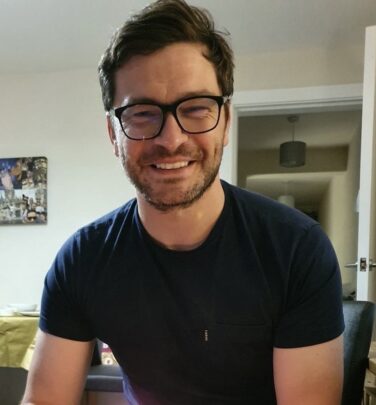 Image of Mike Harrison wearing dark rimmed glasses and a black t-shirt smiling directly at the camera.