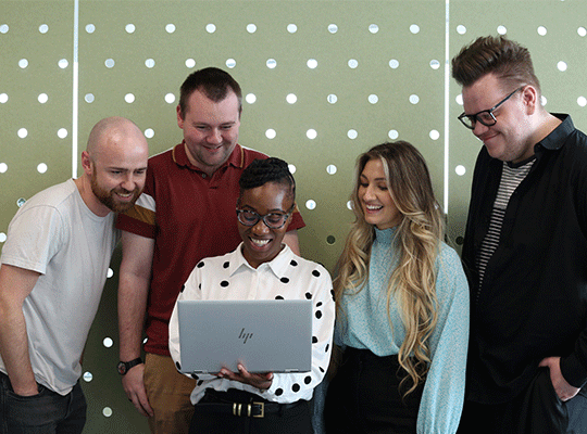 A group of colleagues gathered around 1 laptop looking at the screen.