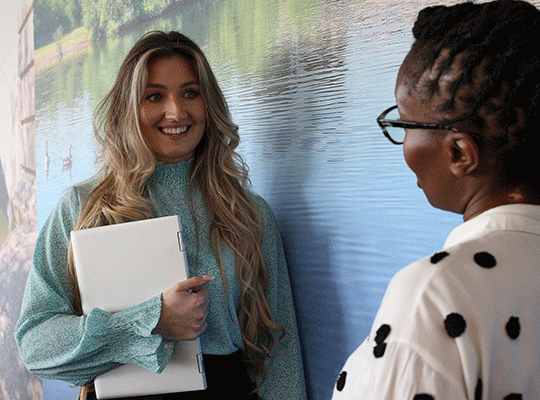 Two colleagues stood chatting in the office with one woman holding a laptop