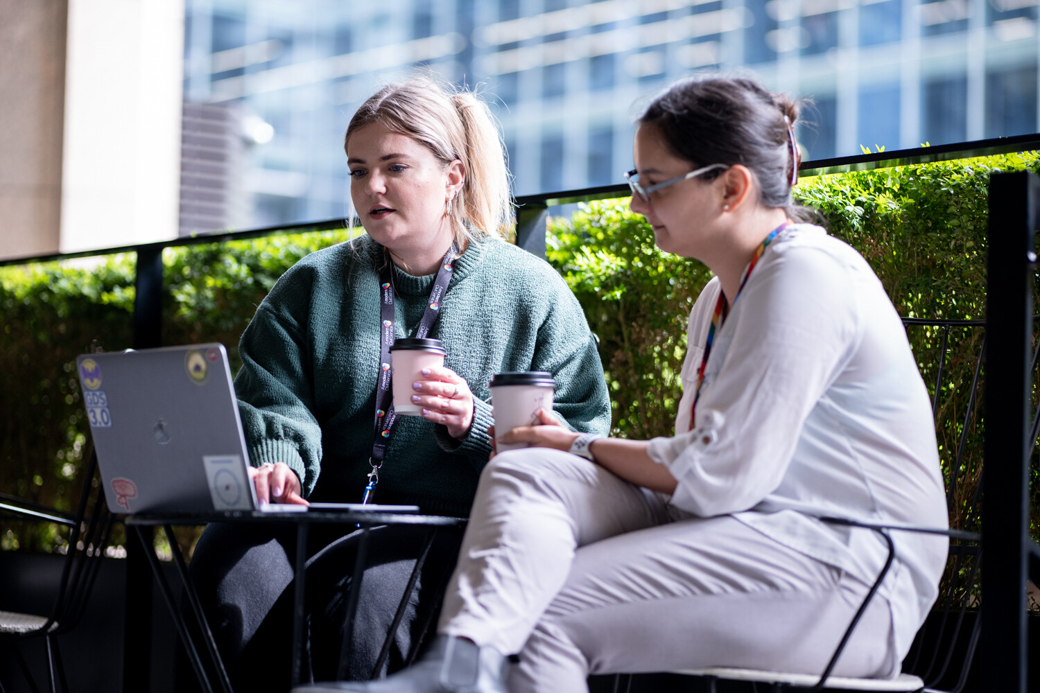Decorative image: two (2) women sitting together outside, working on a laptop and drinking coffee