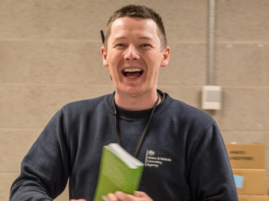 Operational Delivery staff member clutching a green book. He is mid-laugh and has a pen tucked behind one ear.
