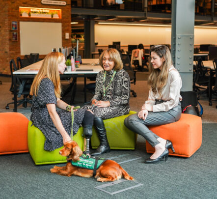 A woman with an assistance dog, and a prosthetic leg, sitting in a colourful break-out area with two colleagues.