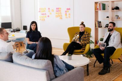 Group of people in an office space sitting and chatting. There is a whiteboard with post-it notes in the background.