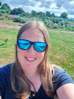 A photo of Hebe Adams, she is outside in a field in the sun, and is wearing mirrored sunglasses.