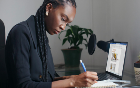 A woman sitting at a desk, writing on a notepad with an open laptop at her elbow