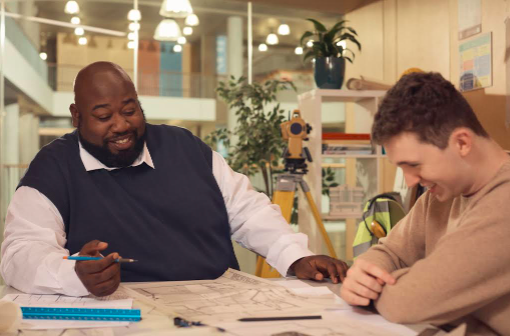 Decorative image: a student meeting with his tutor. They are both men and are sitting at a table strewn with paperwork, both are laughing and look relaxed.