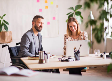 A man and a woman are sitting at a desk, looking at a laptop, the man is a wheelchair user