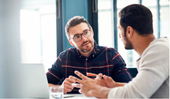 Decorative image: Two men sitting at a desk, having an engaging discussion. One is facing the camera, the other is facing away.