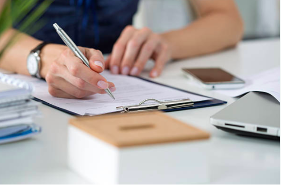Decorative image: A close up of a woman's hands, she's filling in paperwork attached to a clipboard. She's sitting down at a desk.