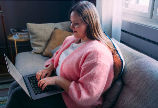 A woman sitting on a sofa, using a laptop on her lap. She's wearing a distinctive pink cardigan