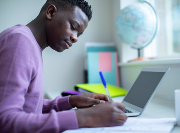 A young man making notes with a pen, with an open laptop beside him.