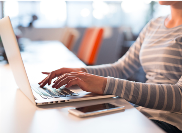 A woman typing on a laptop, with her mobile phone beside her. Her face is not visible, only her neck and torso.